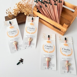 May include: A wooden crate filled with pencils, each with a black tip and a pink body. Several pencils are packaged with a white card featuring a graduation cap graphic and the name "Christina". The cards also include text in Italian.