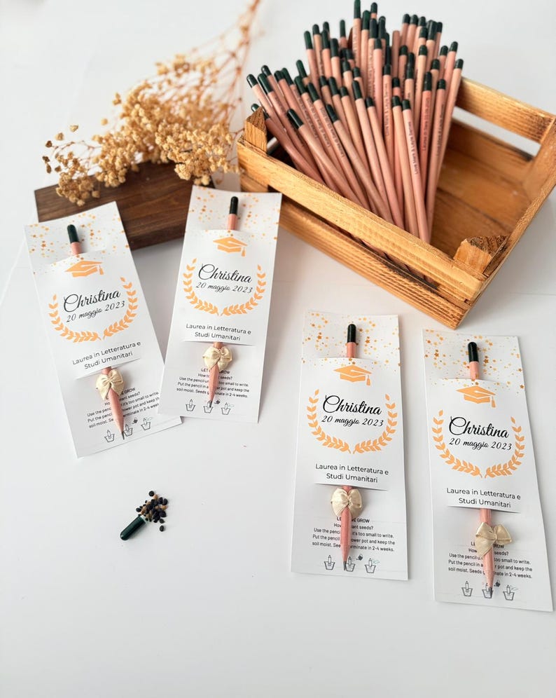 May include: A wooden crate filled with pencils, each with a black tip and a pink body. Several pencils are packaged with a white card featuring a graduation cap graphic and the name "Christina". The cards also include text in Italian.
