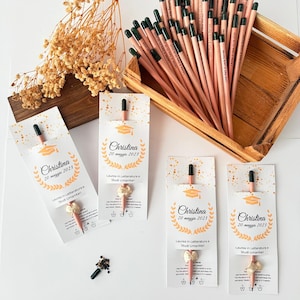 May include: A collection of pencils with green tips and personalized text, displayed in a wooden tray. Several pencils are packaged with cards that read "Christina 20 maggio 2023" and "Laurea in Letteratura e Studi Umanitari."
