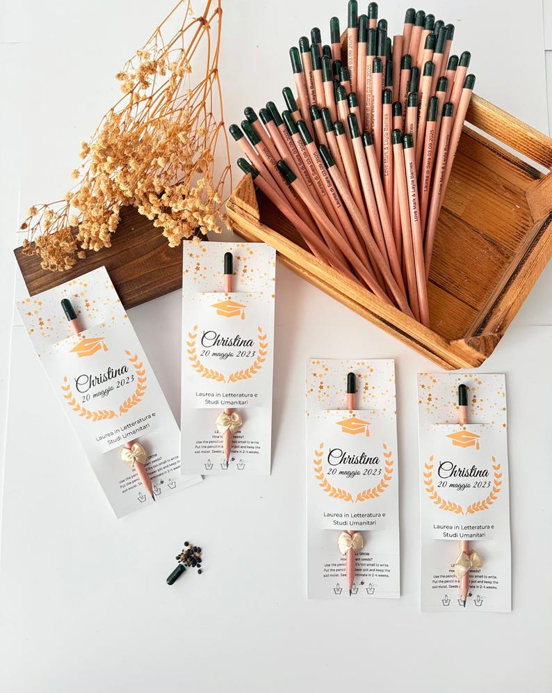 May include: A collection of pencils with green tips and personalized text, displayed in a wooden tray. Several pencils are packaged with cards that read "Christina 20 maggio 2023" and "Laurea in Letteratura e Studi Umanitari."