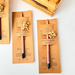 May include: A collection of personalized pencils with attached cards. Each card is tan with the names "Stefano & Francesca" and the date "26.12.2025" printed on it. The pencils are pink with black tips and small dried flowers.