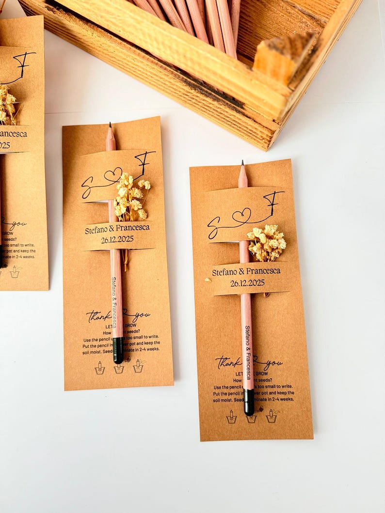 May include: A collection of personalized pencils with attached cards. Each card is tan with the names "Stefano & Francesca" and the date "26.12.2025" printed on it. The pencils are pink with black tips and small dried flowers.