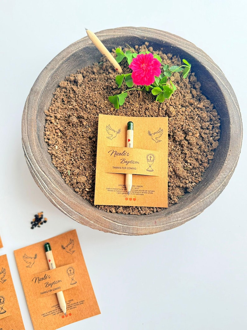 May include: A wooden bowl containing soil, a vibrant pink flower, and a small pencil. A card with the text "Nicole's Baptism" and a pencil is placed in the soil. The card features illustrations of doves and a chalice.