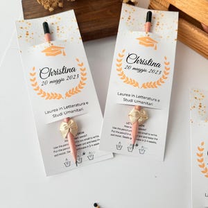 May include: Two white paper packets with a pencil and bow, personalized with the name "Christina" and the date "20 maggio 2023". The packets have a graduation cap graphic and text in Italian.