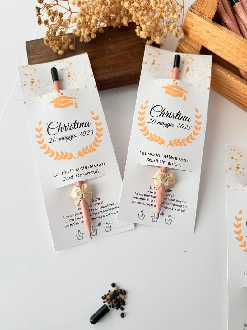 May include: Two white paper packets with a pencil and bow, personalized with the name "Christina" and the date "20 maggio 2023". The packets have a graduation cap graphic and text in Italian.