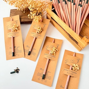 May include: Pink pencils with green tips are displayed with small kraft paper cards. Each card has a small floral arrangement and a tag with a name and date. A wooden box holds more pencils. The pencils are for a wedding.