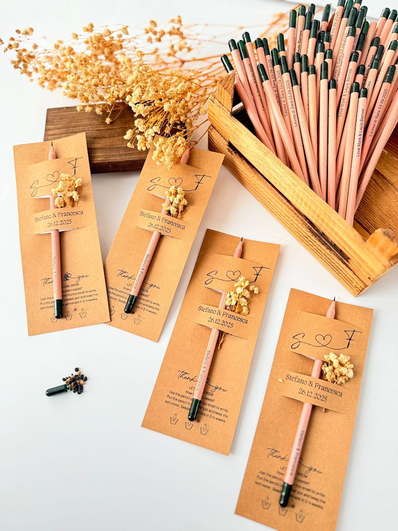 May include: Pink pencils with green tips are displayed with small kraft paper cards. Each card has a small floral arrangement and a tag with a name and date. A wooden box holds more pencils. The pencils are for a wedding.