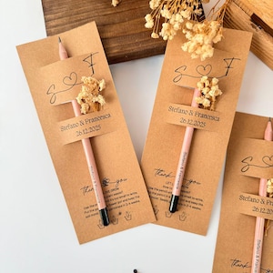 May include: Kraft paper wedding favors with pink pencils, dried flowers, and personalized tags. Each tag reads "Stefano & Francesca 26.12.2025" and includes instructions for planting seeds. A charming and unique wedding favor.