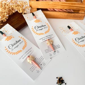 May include: A graduation-themed gift set featuring pencils with seed-embedded tips. Each pencil is packaged in a white card with the name "Christina" and the date "20 maggio 2023", along with a graduation cap graphic and text in Italian.