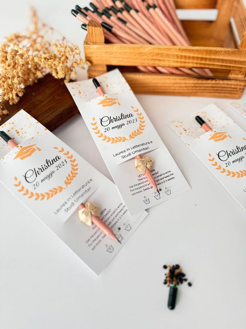 May include: A graduation-themed gift set featuring pencils with seed-embedded tips. Each pencil is packaged in a white card with the name "Christina" and the date "20 maggio 2023", along with a graduation cap graphic and text in Italian.