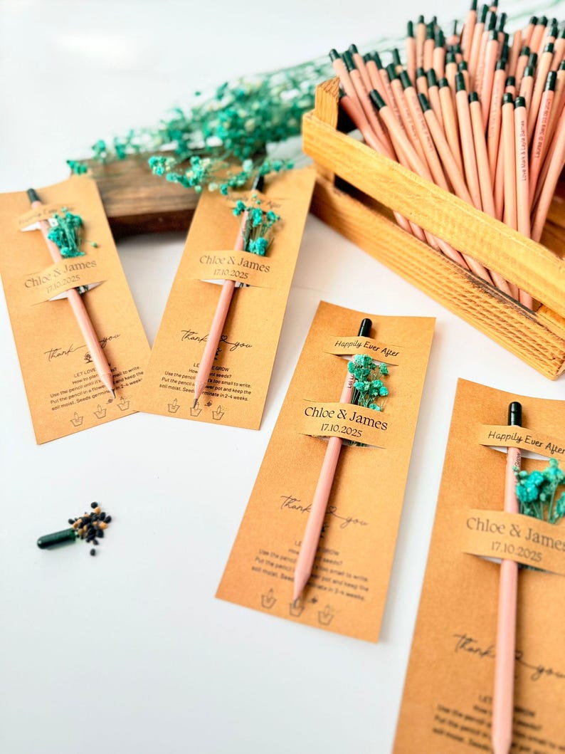 Pu&ograve; includere: Matite rosa con punte nere, attaccate a etichette di carta kraft decorate con piccoli fiori blu. Le etichette riportano "Chloe & James 17.10.2025" e "Happily Ever After". Una cassa di legno contiene altre matite.
