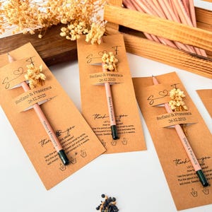 May include: A collection of pencils with attached seed packets, arranged in a wooden crate and on a white surface. Each pencil is adorned with a small floral accent and a tag that reads "Stefano & Francesca 26.12.2025". The pencils are light pink with dark green tips.