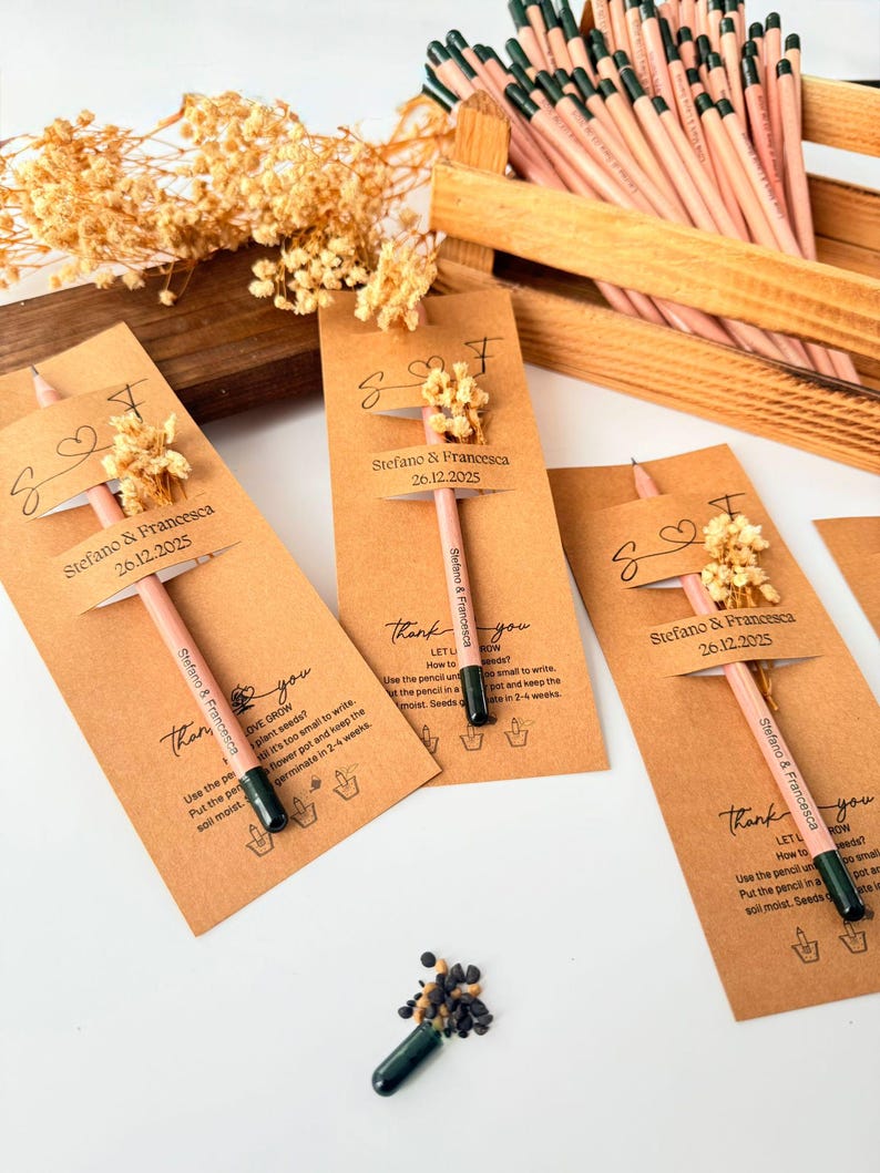 May include: A collection of pencils with attached seed packets, arranged in a wooden crate and on a white surface. Each pencil is adorned with a small floral accent and a tag that reads "Stefano & Francesca 26.12.2025". The pencils are light pink with dark green tips.