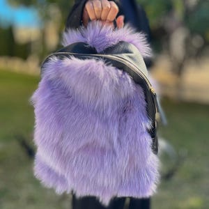 Può includere: Uno zaino nero con un'ampia parte anteriore in pelliccia soffice color lavanda. Lo zaino ha una chiusura a cerniera e una parte superiore in pelle nera. La borsa è tenuta da una persona.