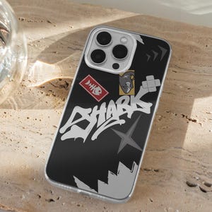 May include: A black phone case with a graphic design featuring the word "SHARK" in white graffiti-style lettering. The case also includes a red and white fishbone icon, a gray star, and a cartoon shark.
