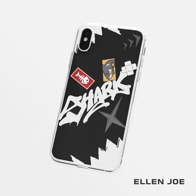 May include: A clear phone case with a black background featuring white graffiti-style text that reads "SHARK." The case has a red rectangular sticker with a fish skeleton and a yellow sticker with a shark illustration. The brand name "ELLEN JOE" is at the bottom.