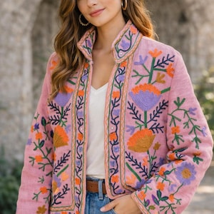 Pode incluir: Casaco rosa bordado com gola alta. O casaco apresenta bordados florais coloridos em laranja, roxo e verde. A modelo usa um top branco e jeans azul.