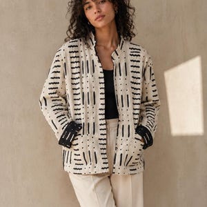 May include: A long-sleeved, cream-colored quilted jacket featuring a black vertical dash pattern, a mandarin collar, and black cuffs. The jacket is worn over a black tank top and paired with loose-fitting, high-waisted cream trousers and black sandals.