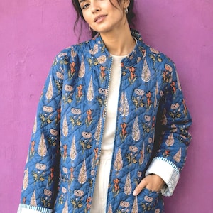 May include: A blue quilted jacket featuring a repeating floral and botanical print in shades of orange, white, and green. The jacket has a mandarin collar, long sleeves with white patterned cuffs, and two front pockets, worn over a white shirt.