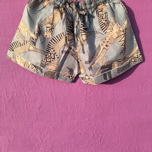 May include: Light blue shorts with an all-over pattern of stylized cat figures in black and beige. The shorts have a drawstring waist and white trim along the edges. The fabric appears to be lightweight.