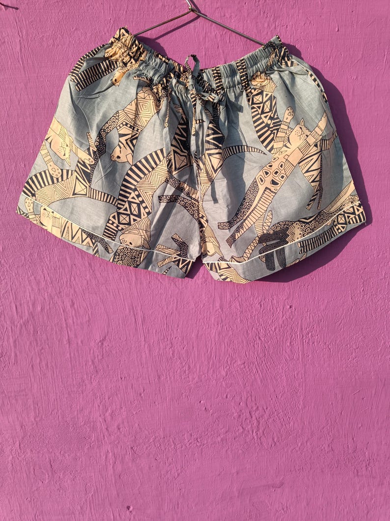 May include: Light blue shorts with an all-over pattern of stylized cat figures in black and beige. The shorts have a drawstring waist and white trim along the edges. The fabric appears to be lightweight.