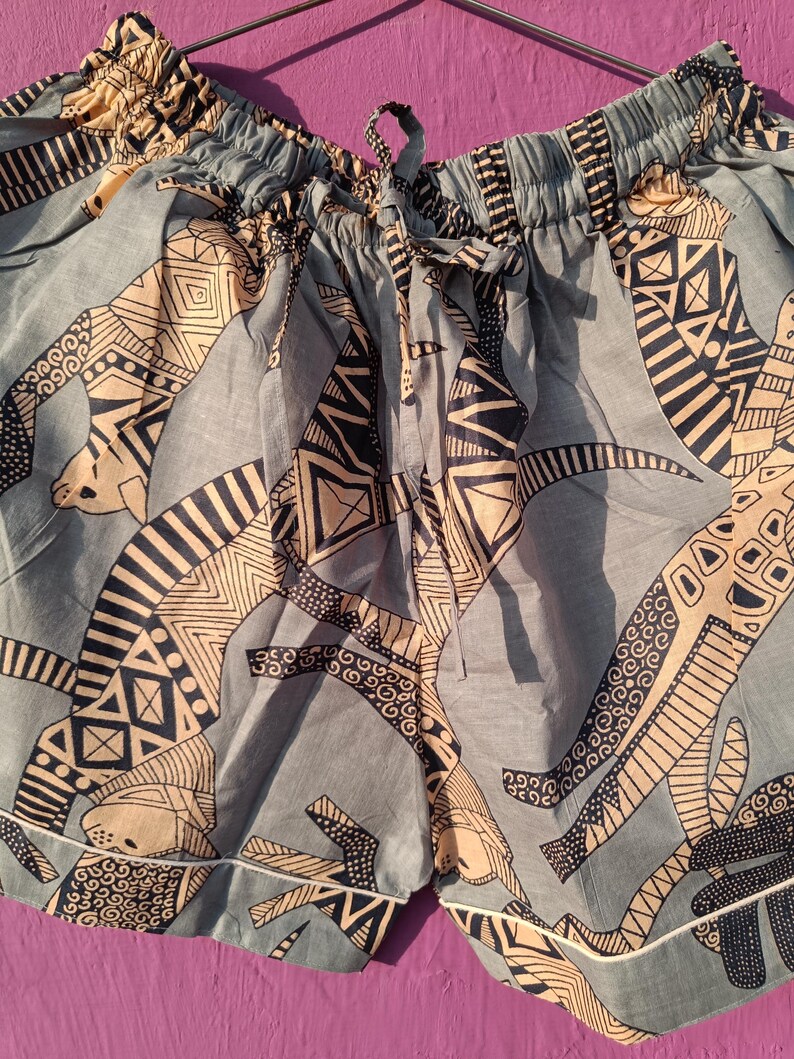 May include: Blue patterned shorts with a drawstring waist and white trim. The shorts feature a repeating design in tan and black. The fabric appears to be lightweight, suitable for warm weather. The shorts are hanging on a metal hanger.
