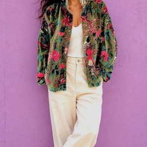 Könnte beinhalten: Eine langärmelige olivgrüne Samtjacke mit einem lebendigen Blumen- und Vogelmuster in Pink und Schwarz, getragen über einem schlichten weißen Tanktop, kombiniert mit einer weiten cremefarbenen Hose und offenen Sandalen.