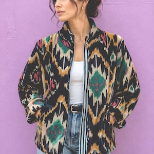 May include: A long-sleeve jacket featuring a black base with a multi-coloured ikat pattern in shades of mustard yellow, teal green, and magenta pink. The jacket has a high collar, a front zip closure, and two side pockets, worn over a white vest top.
