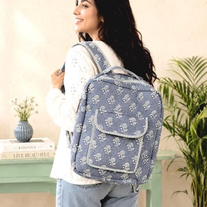 Puede incluir: Una mochila azul acolchada con un estampado floral blanco de bloques. La bolsa incluye un asa superior, correas ajustables para los hombros y un bolsillo frontal con cierre de solapa. La tela tiene una apariencia texturizada y cosida en todo el diseño.