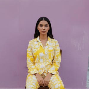 May include: Yellow pajama set with a white floral pattern. The long-sleeved top has a collared neckline and a chest pocket. The matching pants complete the set. The person is sitting on a wooden chair.