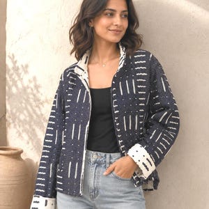 Könnte beinhalten: Eine dunkel marineblaue Steppjacke mit einem weißen, geometrischen, von Mudcloth inspirierten Muster, einem Stehkragen, langen Ärmeln mit umgeschlagenen Manschetten und einem offenen Design, getragen über einem schwarzen Top und hellblauer Jeans.