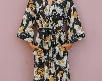 May include: A black robe with a crane pattern in white and gold. The robe has a tie belt, pockets, and white trim. The model is wearing the robe. The background is a solid pink color.