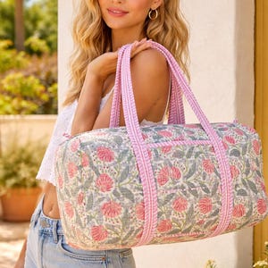 May include: A floral print duffel bag with pink accents. The bag features a pattern of pink flowers and green leaves on a light grey background. Pink striped trim and handles complete the design. The bag is approximately 50 cm long.