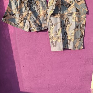 May include: A light blue pajama set with a matching button-down shirt and shorts. The set features a repeating pattern of abstract figures in beige and black. The shorts have a drawstring waist and the shirt has a collared neckline.