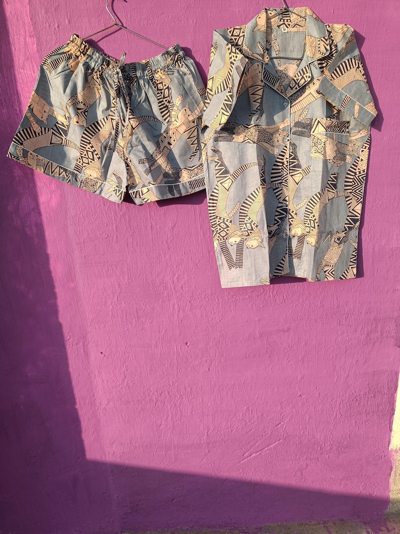 May include: A light blue pajama set with a matching button-down shirt and shorts. The set features a repeating pattern of abstract figures in beige and black. The shorts have a drawstring waist and the shirt has a collared neckline.