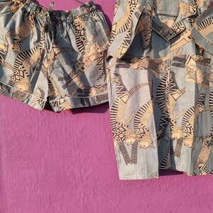 May include: A light blue pajama set with a repeating pattern of stylized figures in black and beige. The set includes a short-sleeved button-up shirt and matching shorts with a drawstring waist, hanging on a metal hanger.
