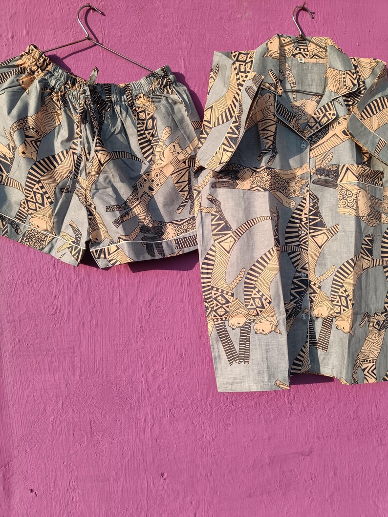 May include: A light blue pajama set with a repeating pattern of stylized figures in black and beige. The set includes a short-sleeved button-up shirt and matching shorts with a drawstring waist, hanging on a metal hanger.