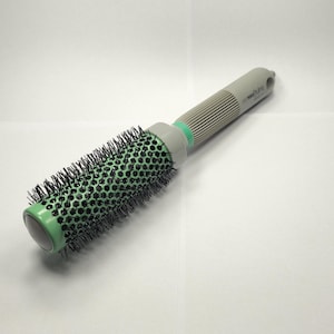 May include: A round hair styling brush with a gray handle and a green barrel. The barrel is covered in black bristles. The handle has a ribbed design for grip and a small hole at the end.