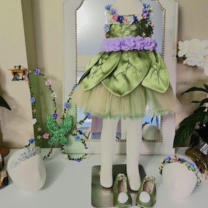 May include: A green dress featuring a bodice adorned with small colorful flowers, a lavender ruffled waist, and a skirt layered with large satin leaf-shaped fabric pieces over a tulle underskirt, accompanied by matching green shoes with white pom-poms.