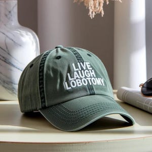 May include: A green baseball cap with the white text "LIVE LAUGH LOBOTOMY". The cap features a curved peak and a dark green stripe. The cap is on a light surface. The cap is a popular fashion accessory.
