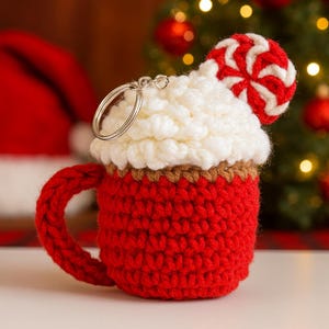 Può includere: Un portachiavi a forma di tazza all'uncinetto in rosso, bianco e marrone. La tazza ha un manico ed è sormontata da filato bianco, che ricorda la panna montata, e una caramella alla menta piperita rossa e bianca. È attaccato un portachiavi argentato.