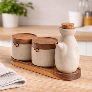 Puede incluir: Un juego de cocina de cerámica con base de madera. El juego incluye dos botes pequeños con tapas de madera y una botella pequeña de cerámica con tapón de corcho. Los elementos de madera son marrones.