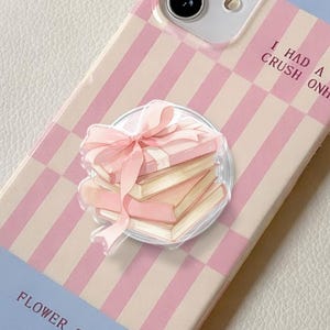 Op de afbeelding: Een lichtblauwe en roze telefoonhoes met een dambordpatroon en een stapel boeken met een roze strik. De hoes heeft de tekst "I HAD A CRUSH ON HIM" en "FLOWER S".