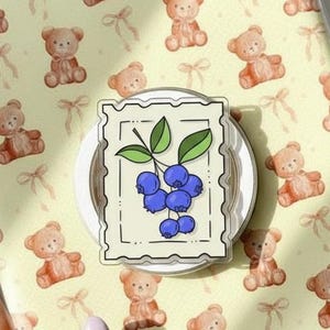 Puede incluir: Una funda transparente para tableta con un fondo amarillo que presenta un patrón de ositos de peluche marrones y lazos. Un pop-socket blanco con una ilustración de arándanos está adherido a la funda.