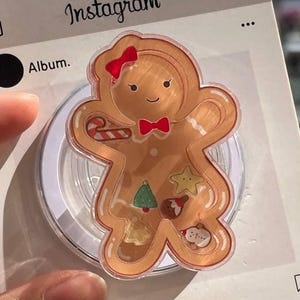 May include: A clear phone grip featuring a gingerbread man design with a red bow and bow tie. The gingerbread man has a candy cane, Christmas tree, star, and other festive shapes inside. The grip is on a white Instagram-themed card.