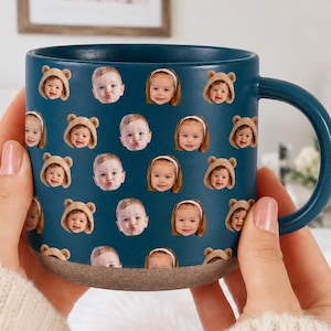 May include: A teal ceramic mug with a pattern of baby faces, some wearing bear hats. The mug has a dark teal handle and a light brown base. The mug is held by a person wearing a cream-coloured sweater.