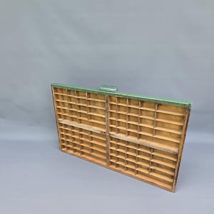 May include: A rectangular wooden printer's drawer featuring a grid of numerous small, open compartments for storage. The frame is painted a muted green, while the interior dividers and base retain a natural, light brown wood finish.