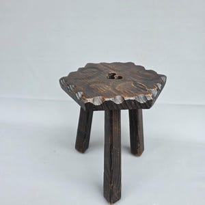 Peut inclure: Un petit tabouret en bois brun foncé doté de trois pieds droits et d'un plateau hexagonal. Les bords de l'assise présentent une texture sculptée en festons, et la surface révèle un grain de bois visible avec une finition sombre et usée.