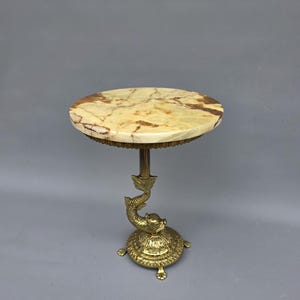 May include: A small, decorative table with a round, marble top featuring brown and cream tones. The table is supported by a gold-coloured metal base with a dolphin design and a circular foot. The table is approximately 51 cm tall.