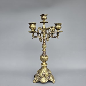 May include: Ornate brass candelabra with five candle holders. The antique candle holder features a detailed design with floral and scrollwork patterns. The base is wide and decorative, and the overall color is a warm gold.